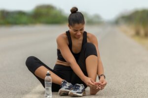 Kobieta w stroju sportowym siedzi na ulicy ze zdjętymi butami i trzyma się za stopę